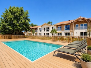 A swimming pool with a wooden deck, surrounded by a wooden fence, and a two-story building with balconies. - The Swallow - Apt for 4 - Shared Pool (Biscarrosse)