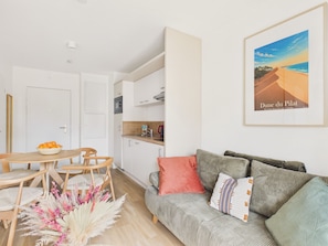 A modern living room with a sofa, a dining table with chairs, a kitchen area, and a framed picture on the wall. - The Swallow - Apt for 4 - Shared Pool (Biscarrosse)