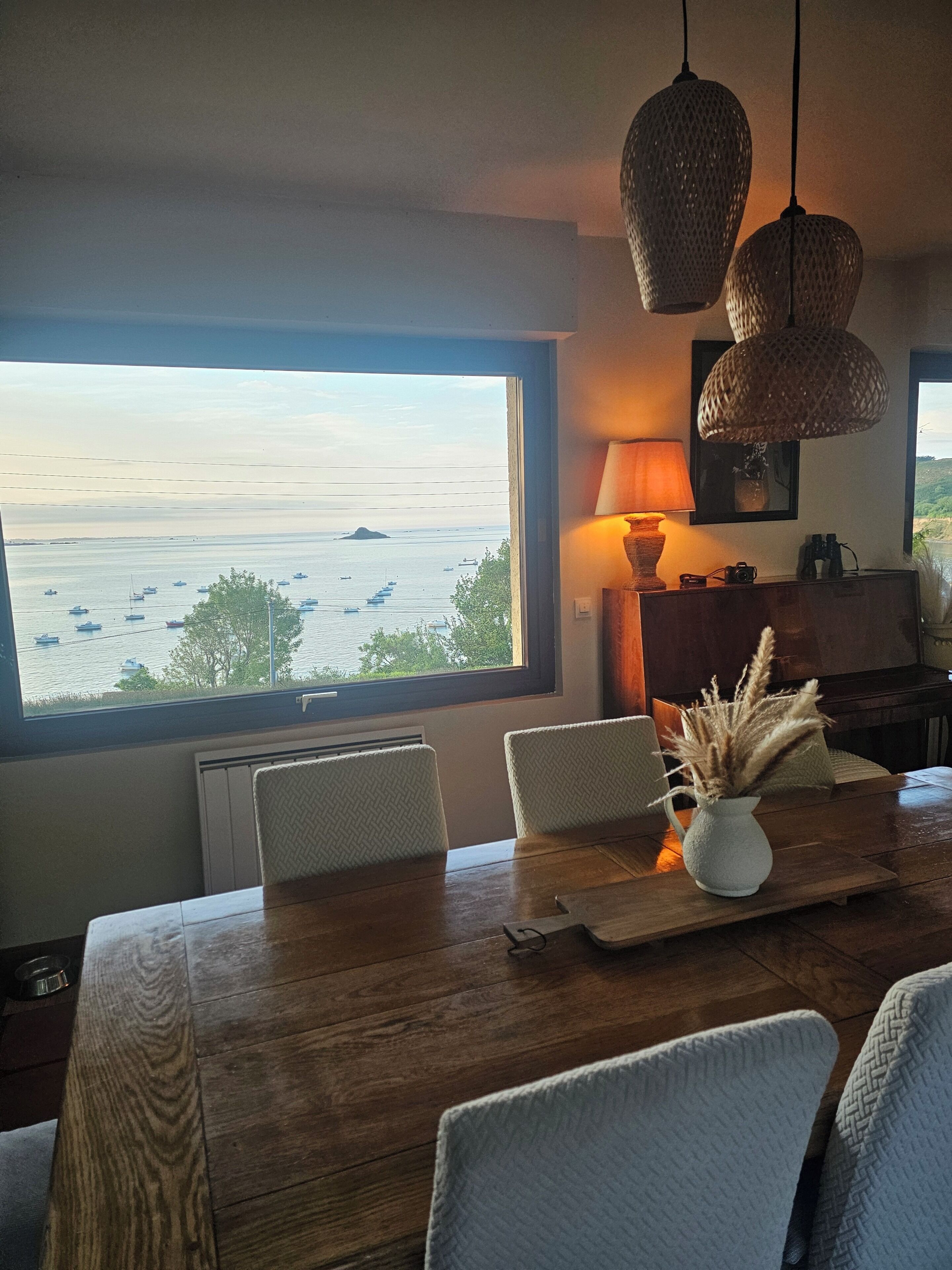 A dining area with a large window overlooking a body of water, a wooden table, and wicker chairs.