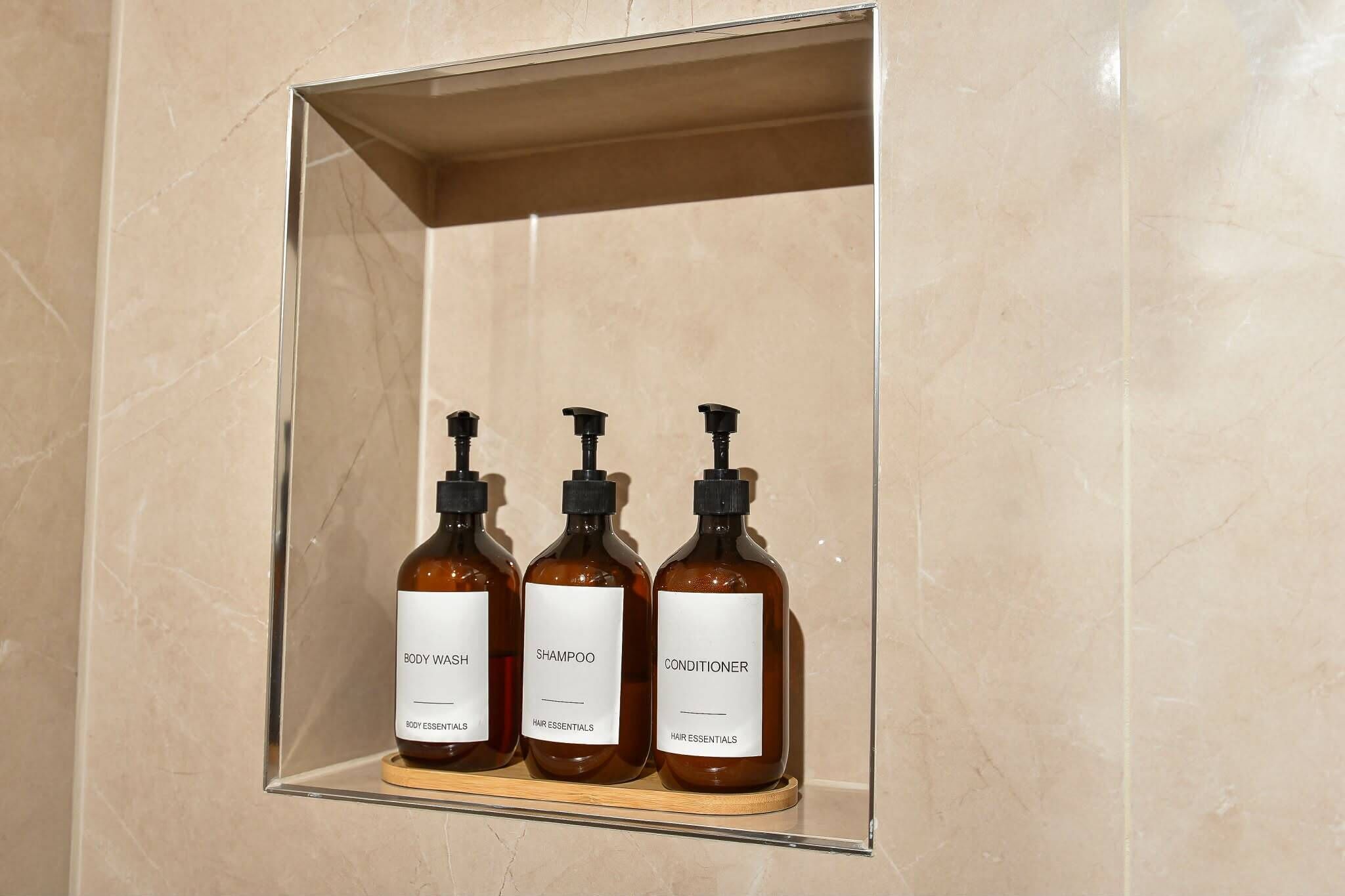 Een badkamer met een marmeren wand, een plank met drie flessen verzorging voor lichaam, shampoo en conditioner.