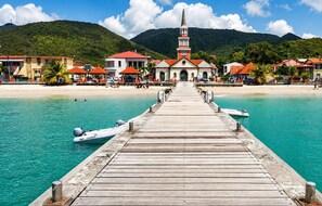 Um cais de madeira que leva a uma praia, com uma igreja e edifícios ao fundo.