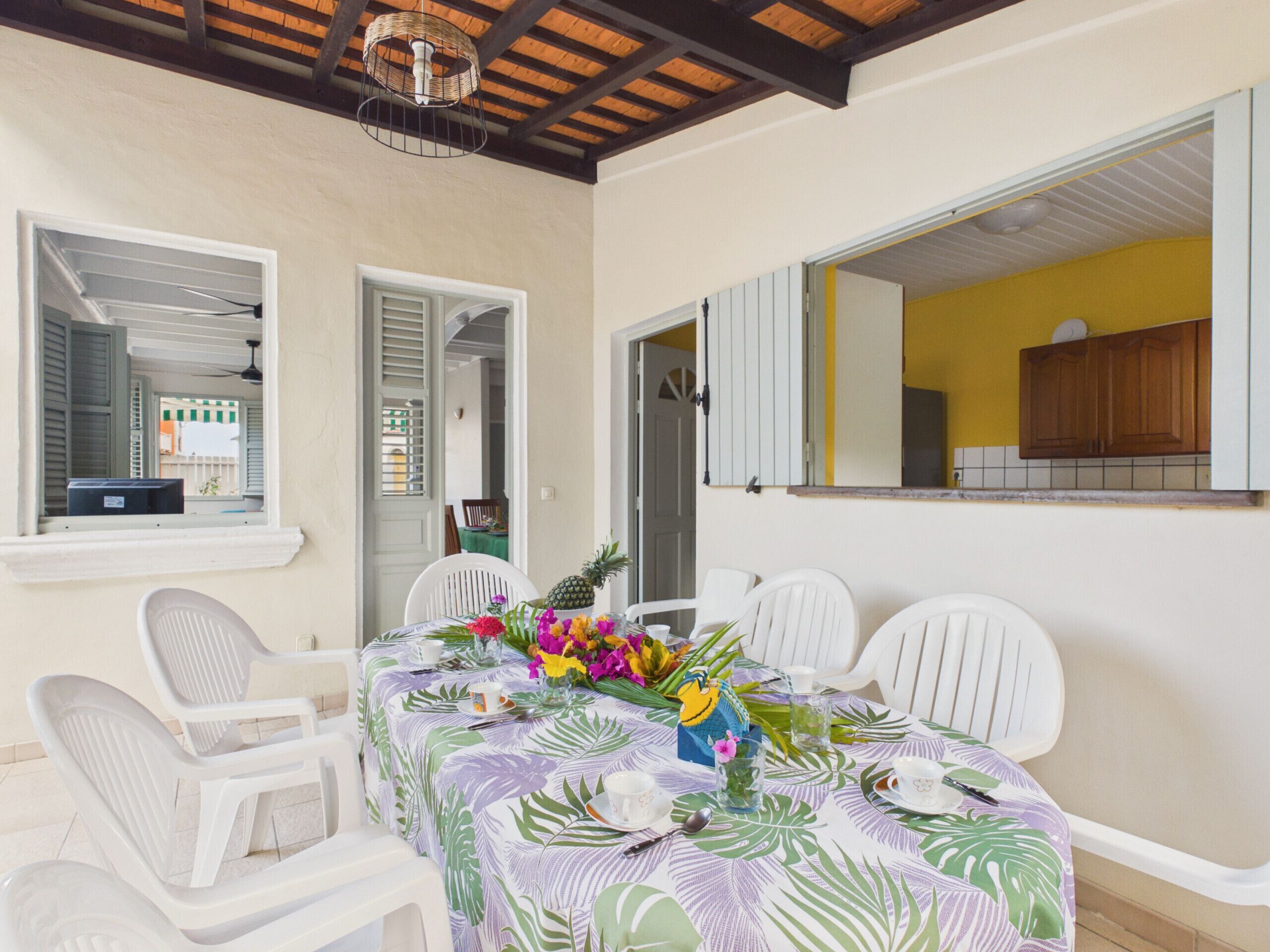 Un coin repas avec une table dressée pour quatre personnes, entourée de chaises blanches, d’une nappe aux motifs tropicaux et d’une pièce maîtresse en forme d’ananas.