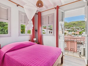 Ein Schlafzimmer mit Bett, einem Fenster mit Rollläden, einem Balkon mit Geländer und Blick auf eine Stadt.