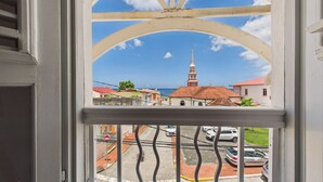 Vista a través d'una finestra amb marc blanc i barana metàl·lica, que dona a un carrer amb cotxes aparcats i edificis amb teulades vermelles.