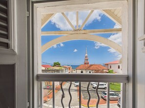 Blick durch ein Fenster mit weißem Rahmen und Metallgitter auf eine Straße mit parkenden Autos und Häusern mit roten Dächern.