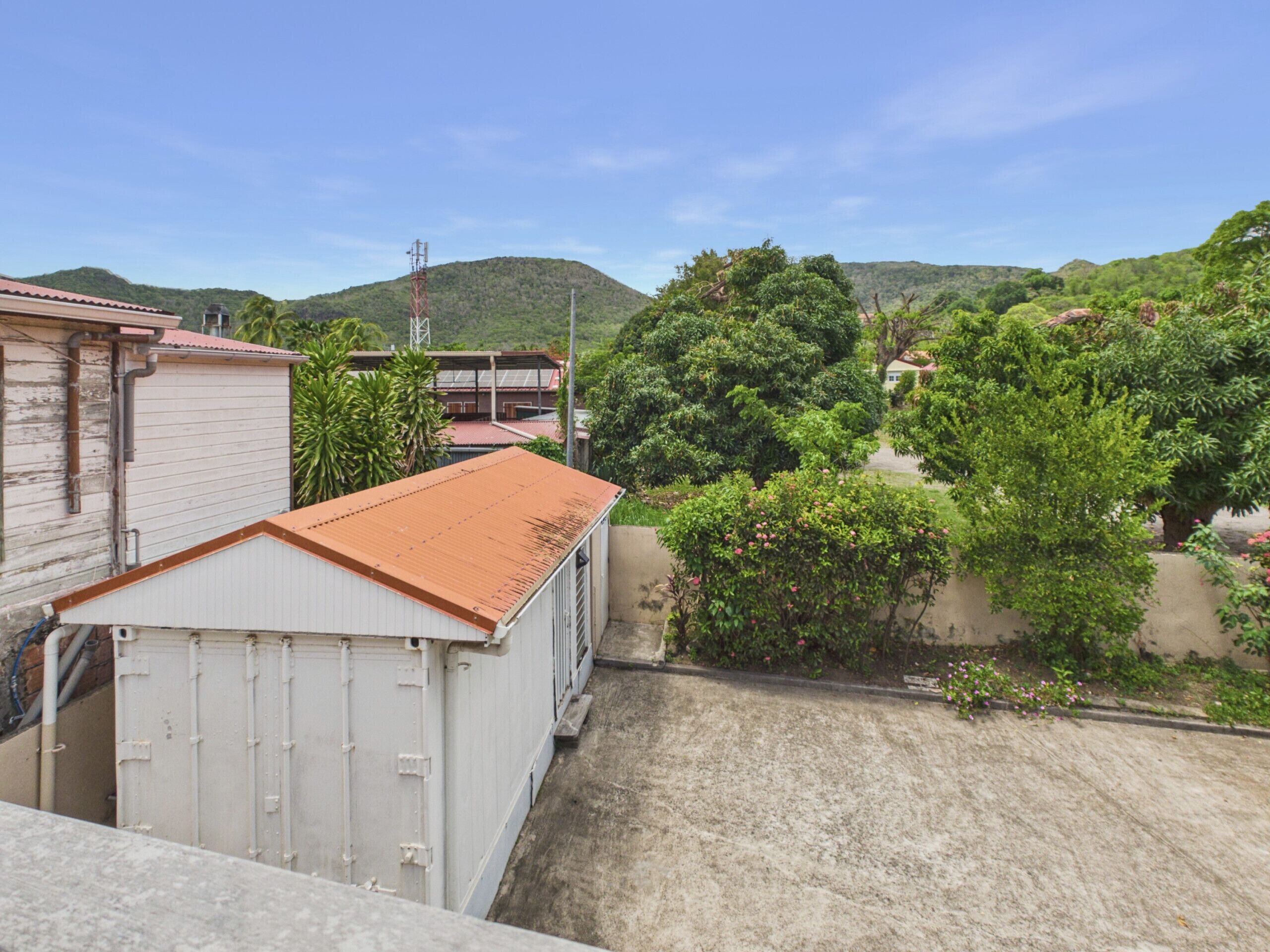 A residential area with a white building with a red roof, surrounded by greenery and hills in the background.