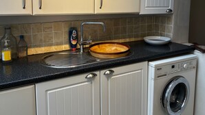 Uma cozinha com armários brancos, uma pia com uma tigela de madeira, uma máquina de lavar e uma garrafa de detergente.