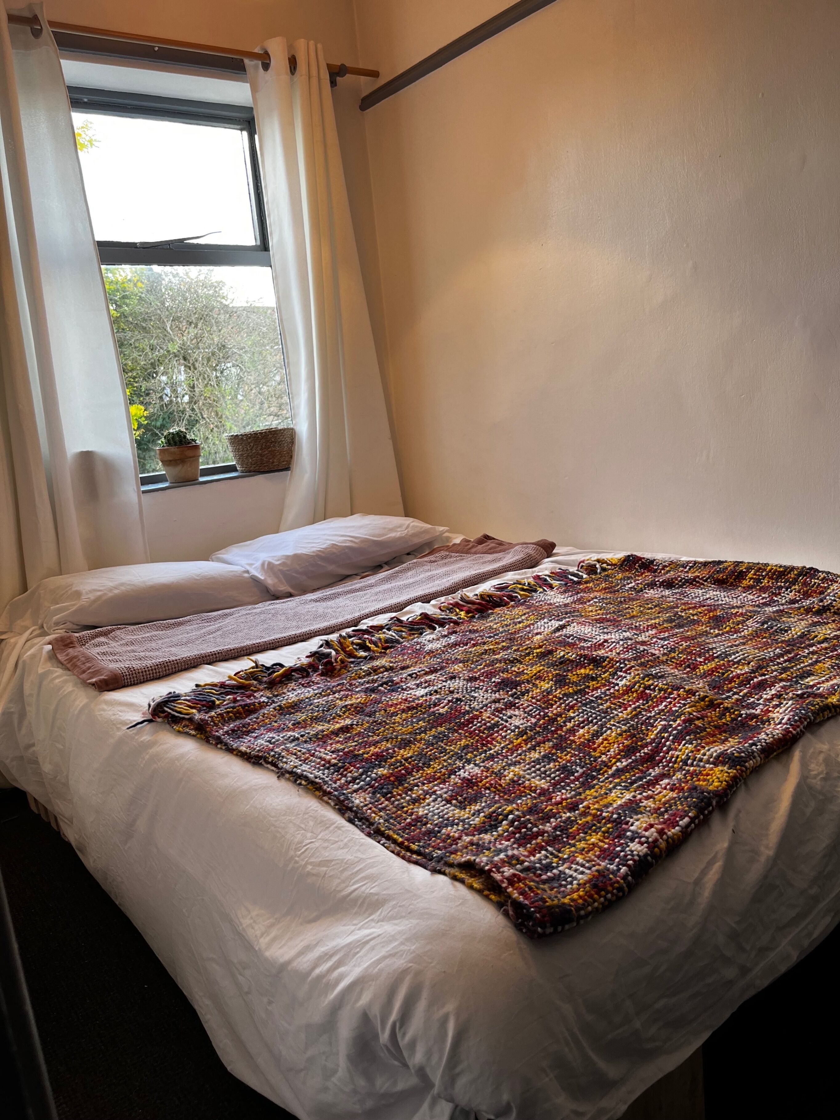 A bed with a white duvet, a colorful woven blanket, and a window with curtains.