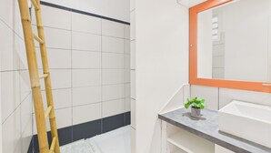 Kylpyhuoneessa on valkoiset laatat, bambusta tehty tikapuu, oranssikehyksinen peili ja pieni ruukkukasvi.