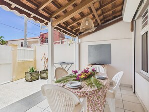 Eine Terrasse im Freien mit Esstisch, umgeben von weißen Stühlen, einer Hängelampe und Blick auf eine Wohngegend.