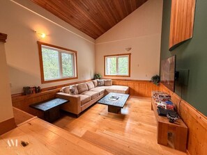 Ein geräumiges Wohnzimmer mit einer großen, L-förmigen Eckcouch, zwei Couchtischen, Holzböden und großen Fenstern mit Blick auf Grünflächen.