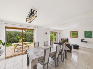 Un coin repas avec une table et des chaises, un téléviseur et une vue sur un jardin, accessible par une porte coulissante.