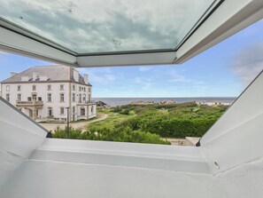 Blick aus einem Fenster auf ein großes Gebäude mit zahlreichen Fenstern und einem Balkon, eine Grünfläche und ein Gewässer in der Ferne.