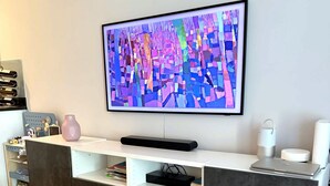 Ein modernes Wohnzimmer mit einem an der Wand befestigten Flachbildfernseher, einem weißen Konsolentisch mit einer rosa Vase und einem grauen Teppich mit Muster.