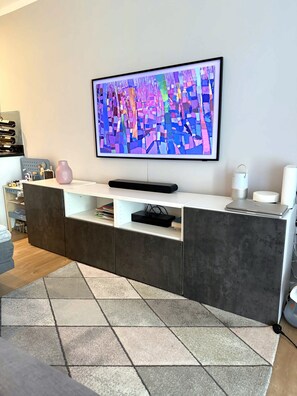 Ein modernes Wohnzimmer mit einem an der Wand befestigten Flachbildfernseher, einem weißen Konsolentisch mit einer rosa Vase und einem grauen Teppich mit Muster.