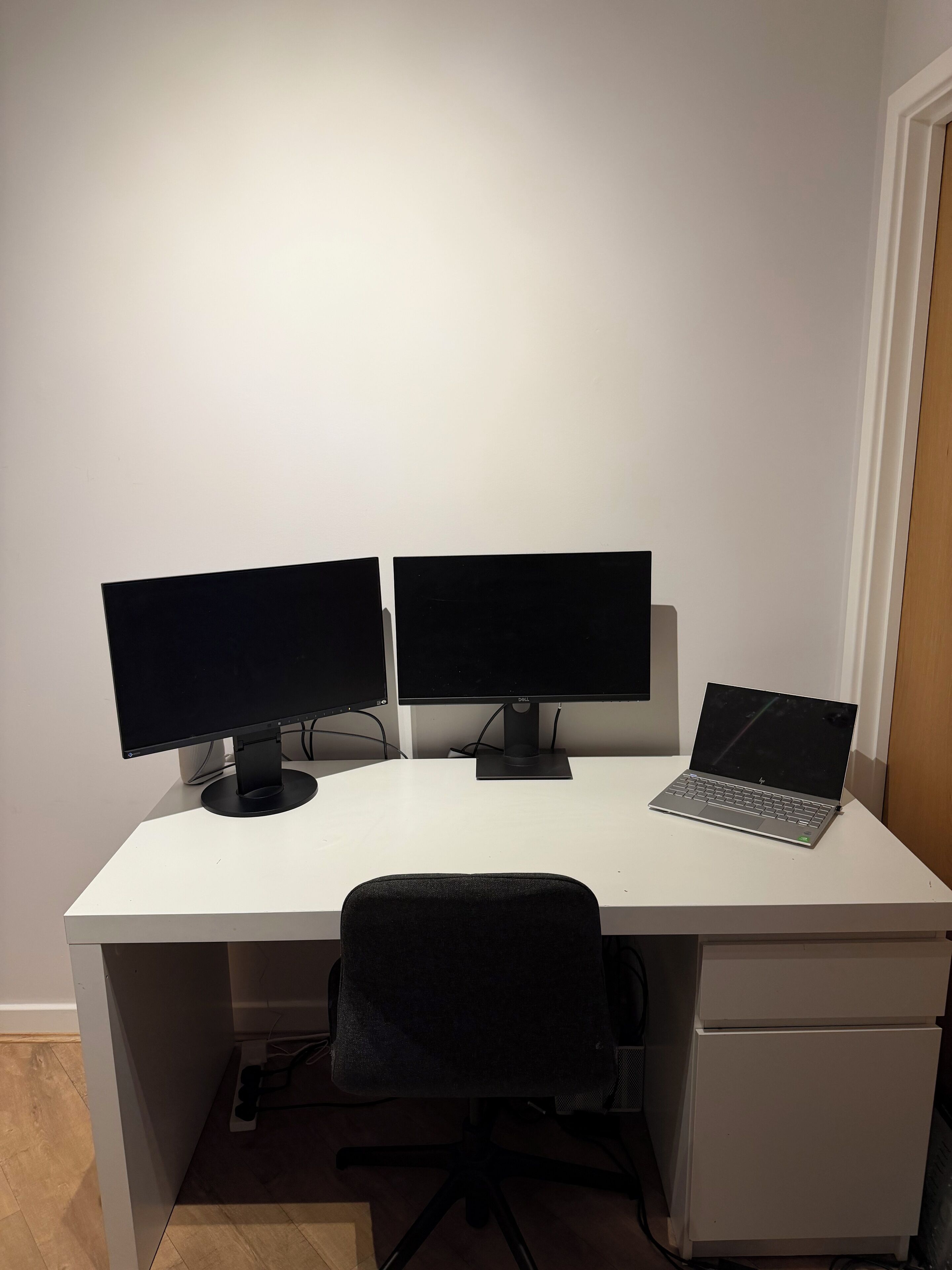 Een wit bureau met twee monitoren en een laptop, een zwarte kantoorstoel en een witte ladekast.
