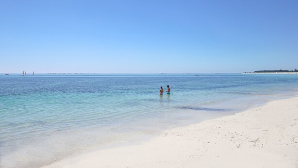 Een strand met helder water en twee mensen die in ondiep water staan.