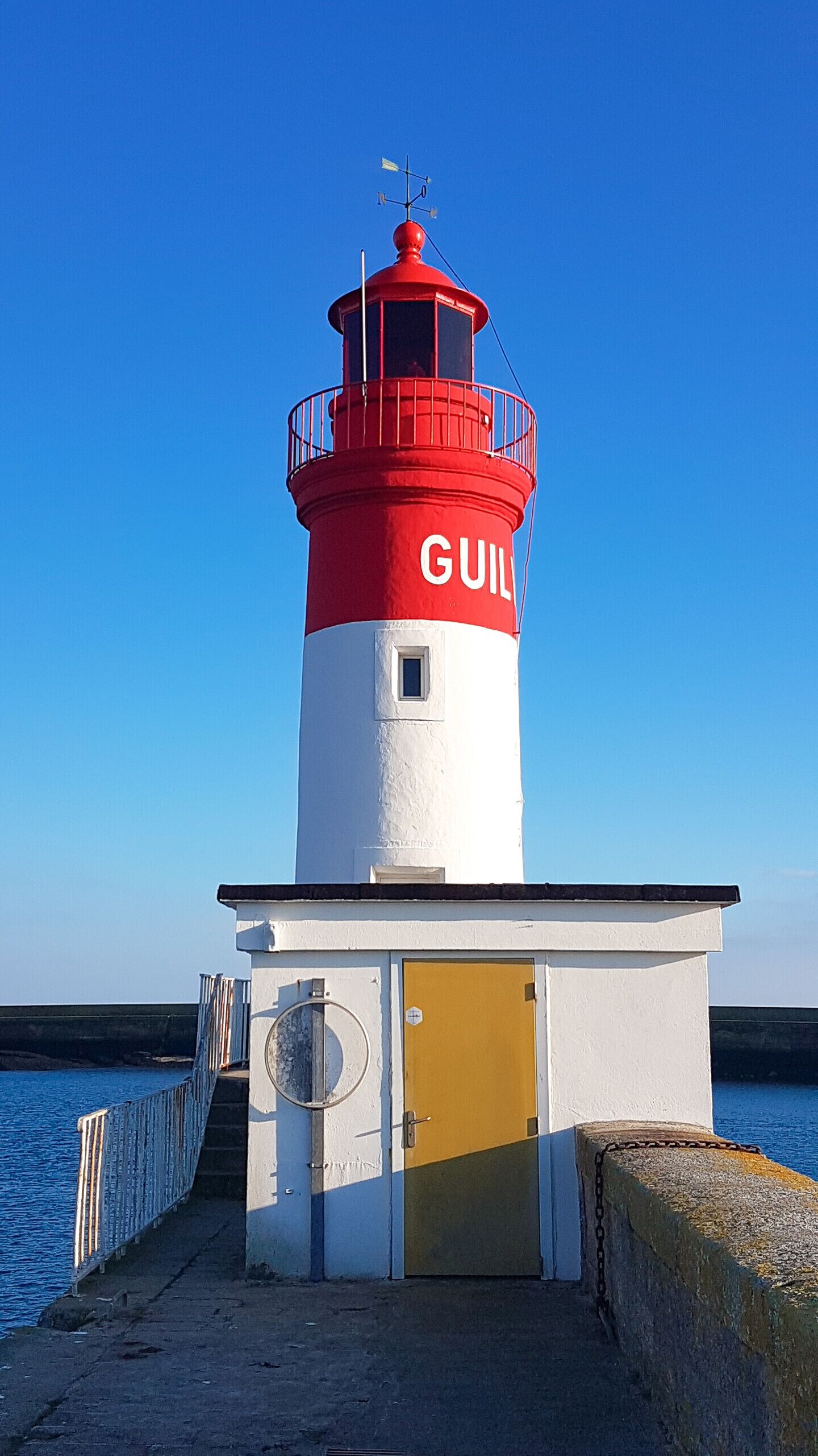 Un phare avec un sommet rouge et un corps blanc, un petit bâtiment d’entrée avec une porte jaune, et une rambarde métallique le long de la jetée.