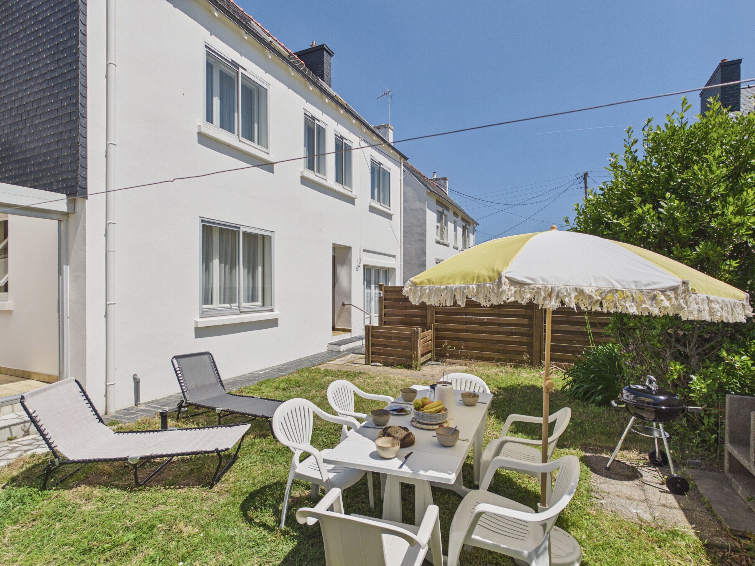 Une maison blanche avec un patio, une table entourée de chaises, un barbecue et un parasol.