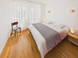 Ein Schlafzimmer mit Holzboden, einem Bett mit weißer Bettwäsche, einem Stuhl, einem Nachttisch mit Lampe und durchsichtigen Vorhängen.