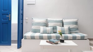 A room with a blue door, a white sofa with striped cushions, a white coffee table with books and a vase of flowers, and a white wall with a light switch. - Folia Bianca-Mi Amo (Imerovigli)
