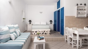A compact living space with a bed, a sofa, a dining table with chairs, a small table with books, and a wall-mounted shelf with bottles. - Folia Bianca-Mi Amo (Imerovigli)