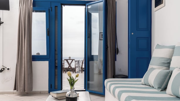 A room with a bed, a white coffee table with a vase, a blue door, and a view of the outside. - Folia Bianca-Mi Amo (Imerovigli)