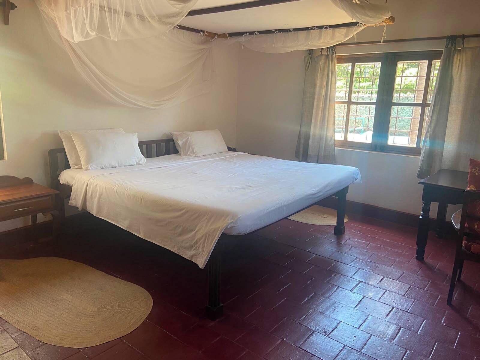 Quarto com cama, mesa de cabeceira de madeira, tapete tecido e janela com cortinas.