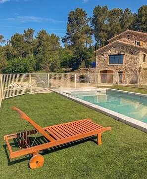 Ein Swimmingpool mit einer Holzliege, umgeben von einem eingezäunten Garten und einem Steinhaus.