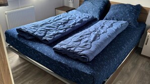 A bed with a blue quilt and pillows, a wooden headboard, a nightstand, and a window with curtains. - Cozy Beach Stay With Climate Control (Kamperland)