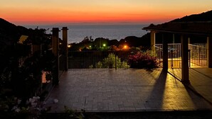 Zonsondergang uitzicht vanaf een betegeld terras met een balustrade, uitkijkend over een wateroppervlak en heuvels in de verte.