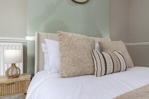 Ein Bett mit weißer Bettwäsche, zwei gemusterte Kissen, ein gewobener Nachttisch und eine Wanduhr.