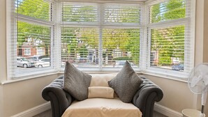 Een zithoek met een comfortabele bank, twee kussens, een ventilator en uitzicht op de straat door grote ramen.