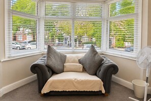 A corner seating area with a cushioned bench, two pillows, a fan, and a view of the street through large windows. - Spacious 3BR Flat Sleeps 7 in Greenford (London)