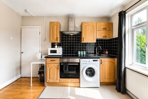 A compact kitchen with wooden cabinets, a microwave, an oven, and a washing machine. - Spacious 3BR Flat Sleeps 7 in Greenford (London)