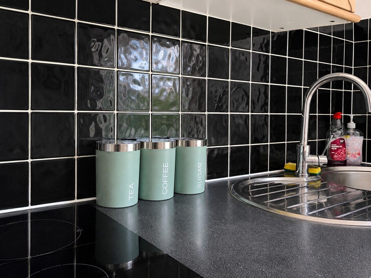 A kitchen with black tiled backsplash, a stainless steel sink, and three teal-colored containers labeled tea, coffee, and sugar.