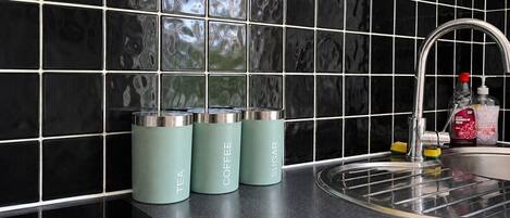 A kitchen with black tiled backsplash, a stainless steel sink, and three teal-colored containers labeled tea, coffee, and sugar.