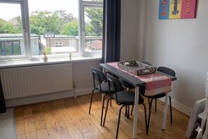 A dining area with a table, chairs, a radiator, and a window with a view of houses and trees. - Spacious 3BR Flat Sleeps 7 in Greenford (London)