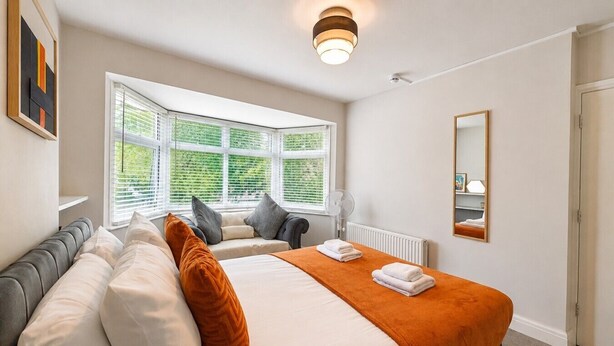 Een hotelkamer met een bed, een raam met jaloezieën, een radiator, een spiegel en een ventilator.