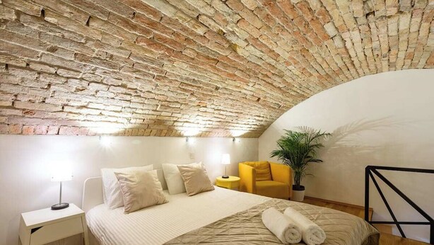 A bedroom with a brick ceiling, a bed with a beige quilt, a nightstand with a lamp, a yellow chair, and a potted plant. - Modern stylish apartment @ Superb central location (R) (Budapest)