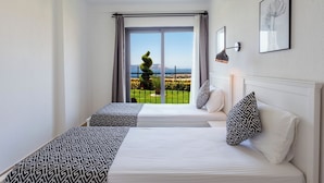 Une chambre d’hôtel avec deux lits, une grande fenêtre donnant sur un paysage verdoyant, un luminaire au plafond et une lampe fixée au mur.
