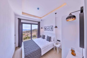 A modern bedroom with a large bed, a bedside table, a nightstand, a window with curtains, and a view of the city. - Lobelya Suites  (Kas)