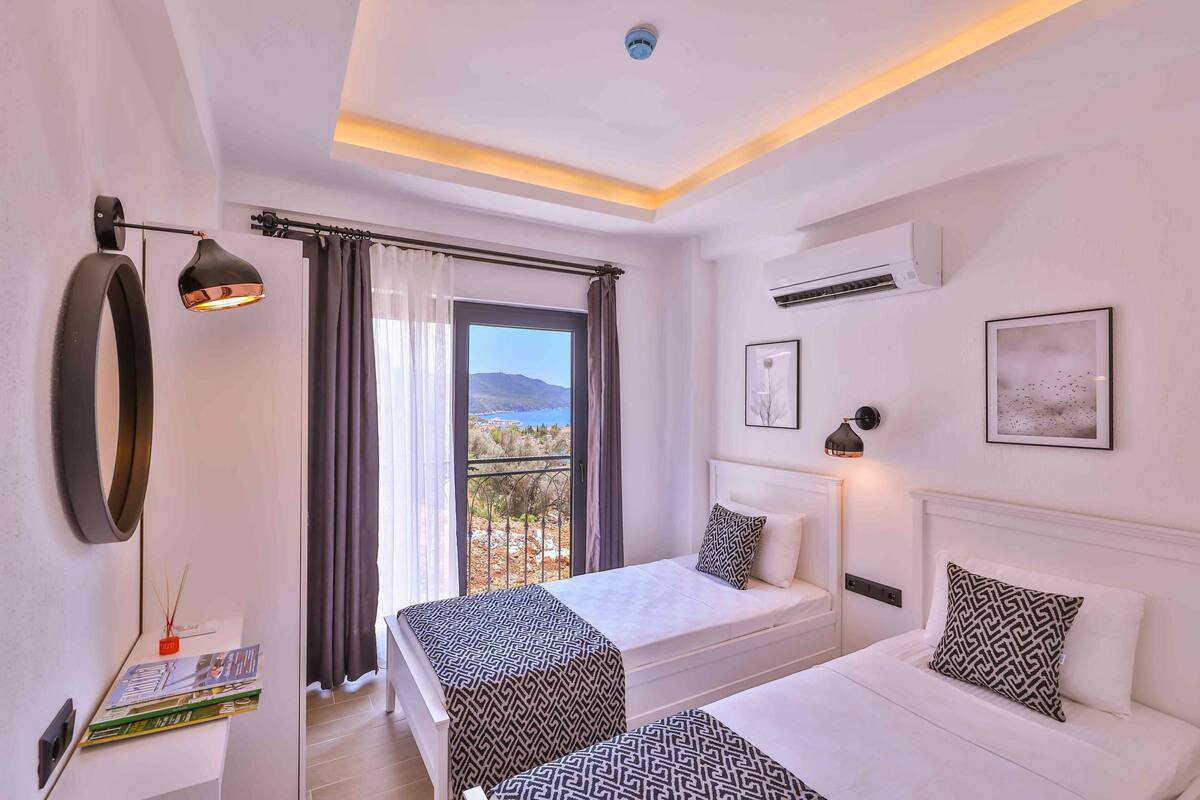 A hotel room with two beds, a balcony with curtains, an air conditioning unit, and a view of the landscape.