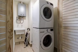 Ein Waschraum mit einer übereinander gestapelten Waschmaschine und einem Trockner, einem weißen Waschbecken und einem Warmwasserbereiter.