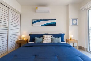 Ein Schlafzimmer mit einem Bett, zwei Nachttischen, einer Wand-Klimaanlage, einem gerahmten Kunstwerk und einem Fenster.
