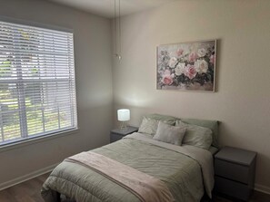 A bedroom with a bed, a nightstand, a lamp, a window with blinds, and a floral painting on the wall.