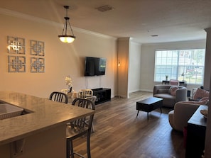 A modern living room with a dining area, a flat-screen TV mounted on the wall, and a large window providing natural light. - Modern Retreat w/ Pool, Bowling Alley & Grills (Oviedo)