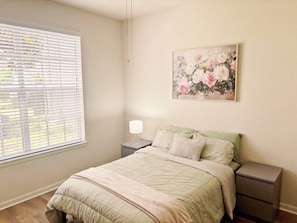 A bedroom with a bed, bedside tables, a lamp, a window with blinds, and a floral painting on the wall. - Modern Retreat w/ Pool, Bowling Alley & Grills (Oviedo)