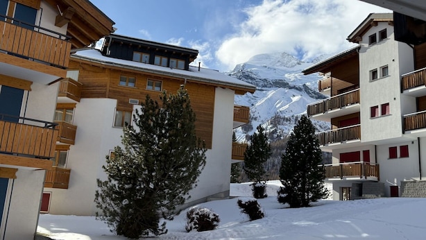 Un patio cubierto de nieve con edificios de madera, árboles nevados y una montaña al fondo.