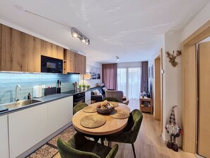 Eine kompakte Küche mit modernen Geräten, ein Essbereich mit rundem Tisch und grünen Stühlen sowie ein gemütlicher Wohnbereich mit Sofa und Vorhängen.